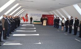 stoi grupa osób, mężczyźni i kobiety, w głębi sali dwaj mężczyźni przemawiają przez mikrofon