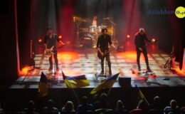 Na scenie stoi trzech muzyków, którzy grają na gitarach, w głębi widać perkusistę, przed muzykami widać publiczność , która trzyma flagi Ukraińskie