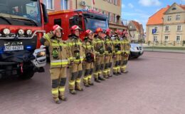 Zdjęcie przedstawia grupę salutujących strażaków na tle strażackich wozów bojowych