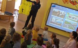 policjantka prezentuje siedzącym przedszkolakom kamizelkę odblaskową