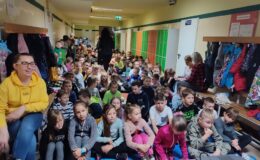 na korytarzu szkolnym, siedzi grupa uczniów i nauczycieli