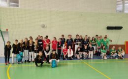 pamiątkowa fotografia zespołów uczestniczących w turnieju baseballu, widać dzieci, dorosłych, którzy stoją i kucają w sali gimnastycznej szkoły