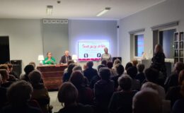 ktorzy Wiesława Szymańska i Marian Czarkowski siedzą przy stoliku, Elżbieta Lenkiewicz stoi, publiczność siedzi na widowni w Miejskiej Bibliotece Publicznej w Działdowie