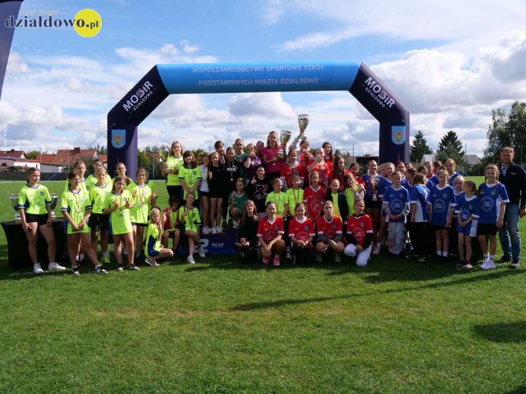 Inauguracja Sportowego Roku Szkolnego 2025/2026 we Współzawodnictwie Szkół Podstawowych w Działdowie stoi grupa osób, młodzież i dorośli. Młodzież stoi na podium, podnosi i trzyma w rękach puchary