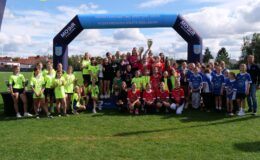 stoi grupa osób, młodzież i dorośli. Młodzież stoi na podium, podnosi i trzyma w rękach puchary