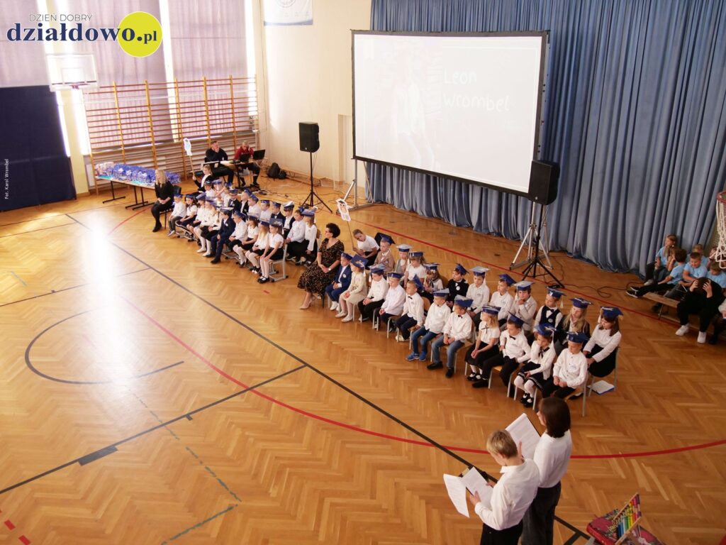 siedzi grupa dzieci i nauczycieli, w sali gimnastycznej Szkoły Podstawowej nr  4  w Działdowie