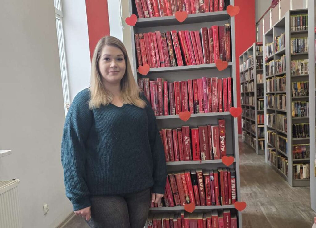 Mielna Matuszewska z Miejskiej Biblioteki Publicznej w Działdowie przy regale z książkowymi walentynkowymi propozycjami