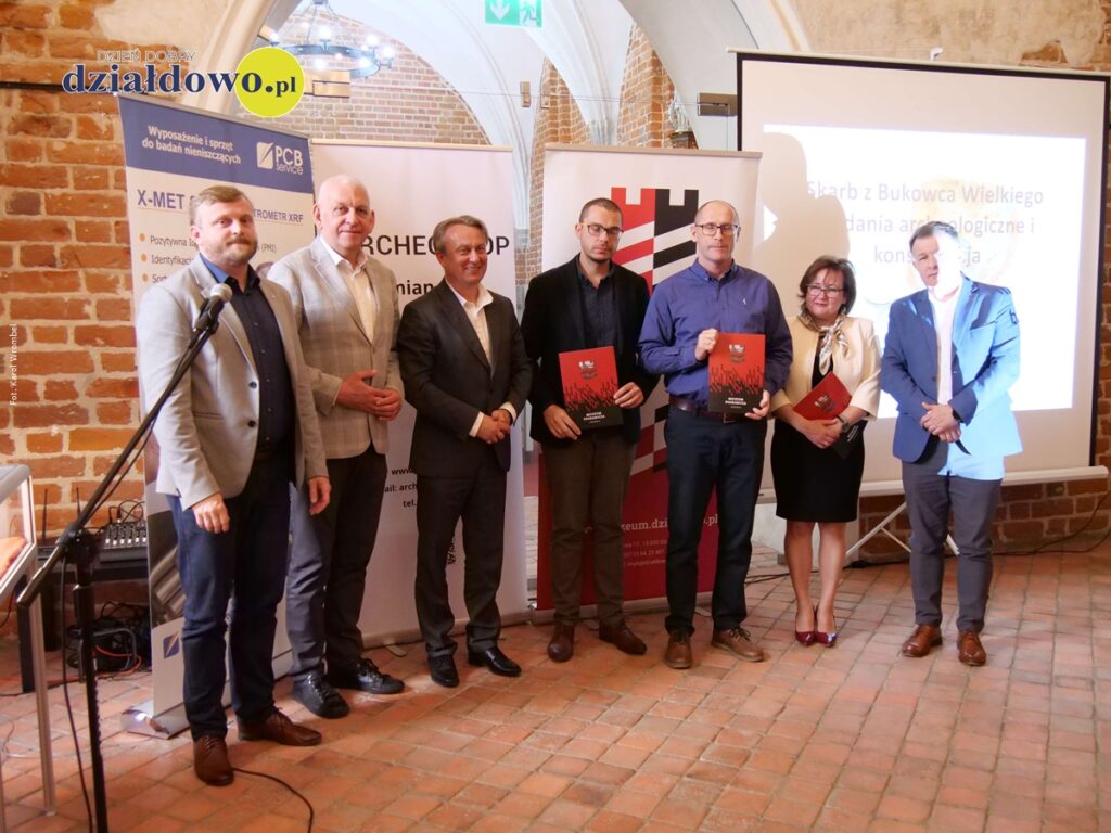 stoją osoby dorosłe podczas konferencji, na której zaprezentowano XVII - wieczne monety po konserwacji, znalezione w Bukowcu Wielkim